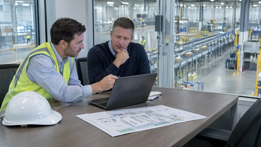 Two men at a conference table beside a glass wall, one pointing at a laptop, with a white hard hat and a layout drawing on the table and a conveyor floor behind them